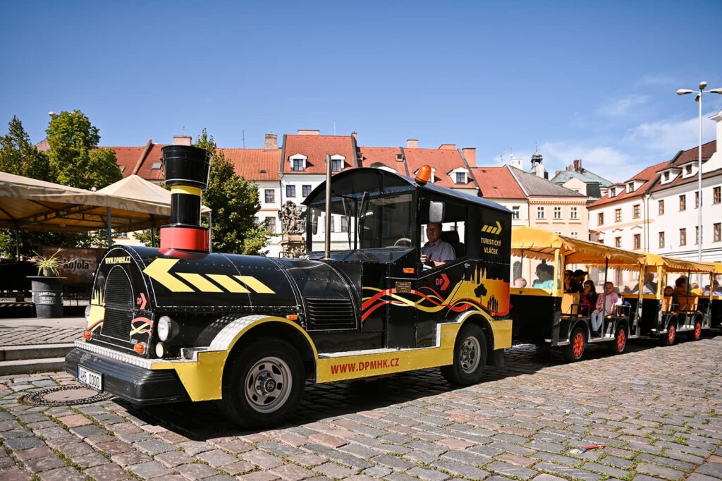 Hradecký turistický vláček s cestujícími projíždí kolem Malého náměstí směrem na Velké náměstí za slunečného počasí
