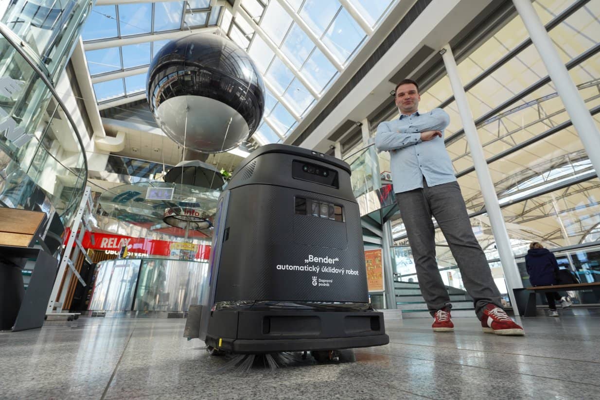 Low-angle view of a black automatic cleaning robot with the text „Bender automatic cleaning robot - Transportation Company“. Next to the robot stands a man with folded arms, wearing a shirt and sneakers. They are in a modern, high-ceilinged glass hall, with a large black decorative sphere suspended in the space and a Relay shop dominating the background.
