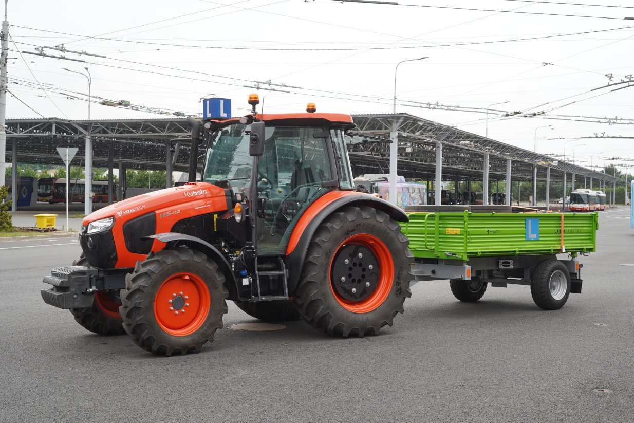 Černo-červený traktor s připojeným zeleným vozíkem stojí v areálu Dopravního podniku. Na vozíku a střeše má značení autoškoly v podobě modré cedule s bílým písmenem L. Za ním je vidět zastřešená plocha se zaparkovanými autobusy.