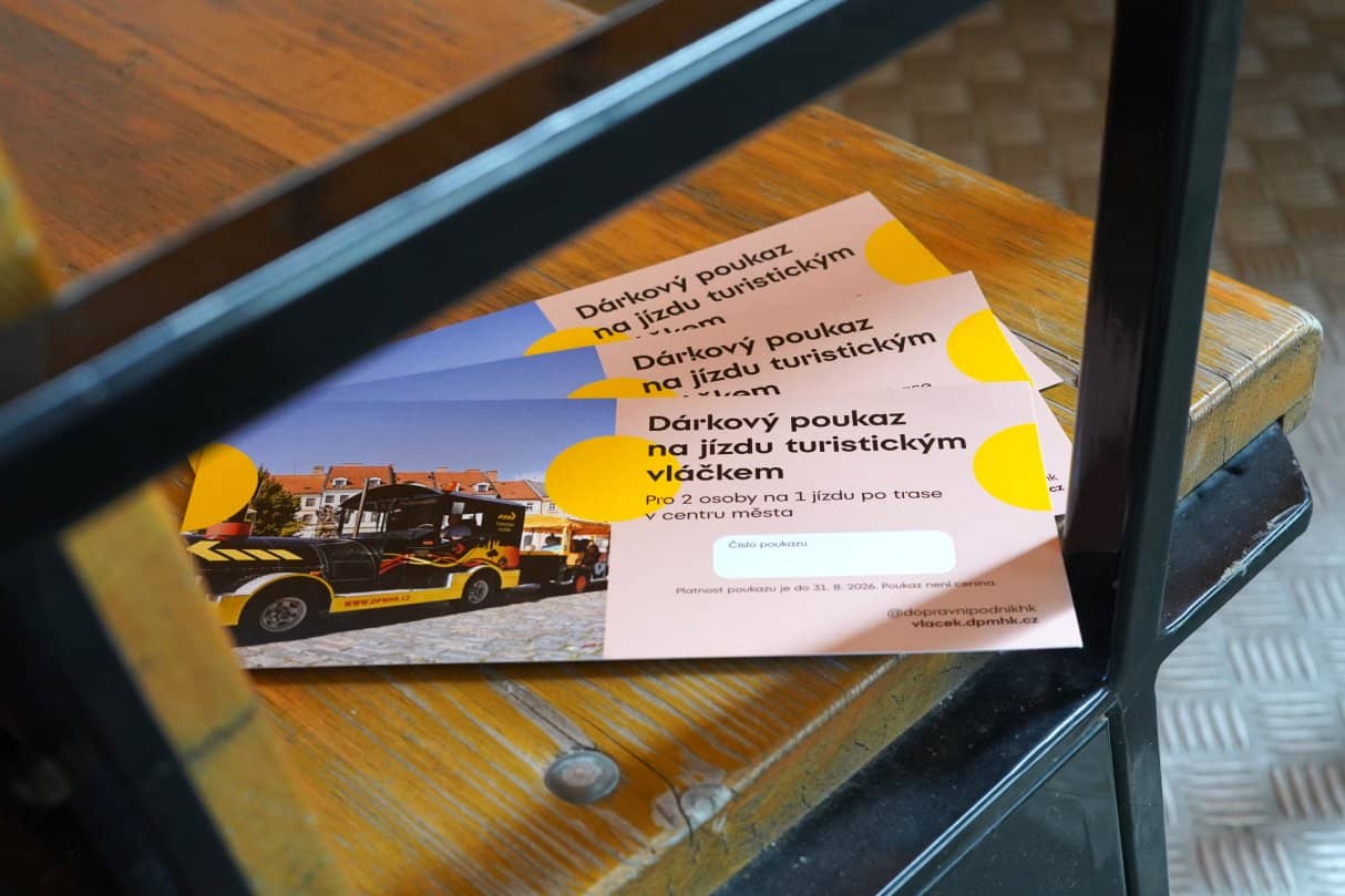 Dárkový poukaz s logem DPMHK a fotografií žluto-černého silničního vláčku na dlážděné ulici v centru města. Poukaz je položen na dřevěné lamelové lavici, má nadpis „Dárkový poukaz na jízdu turistickým vláčkem“ a pod ním text „Pro 2 osoby na 1 jízdu po trase v centru města“. Pod ním jsou další tři stejné poukazy. Scéna je orámována tmavou kovovou konstrukcí a v pozadí je vidět podlaha s mřížkou.