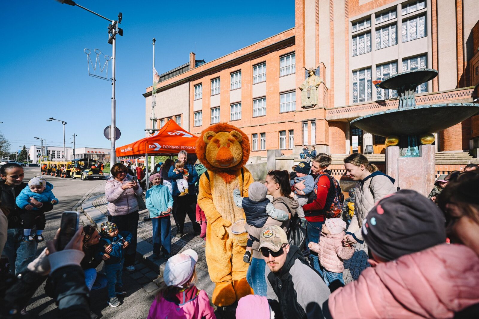 Maskot lva Gustíka baví rodiče s dětmi na prostranství před budovou Muzea východní Čech a fontánou, v pozadí stojí žlutočerný turistický vláček.