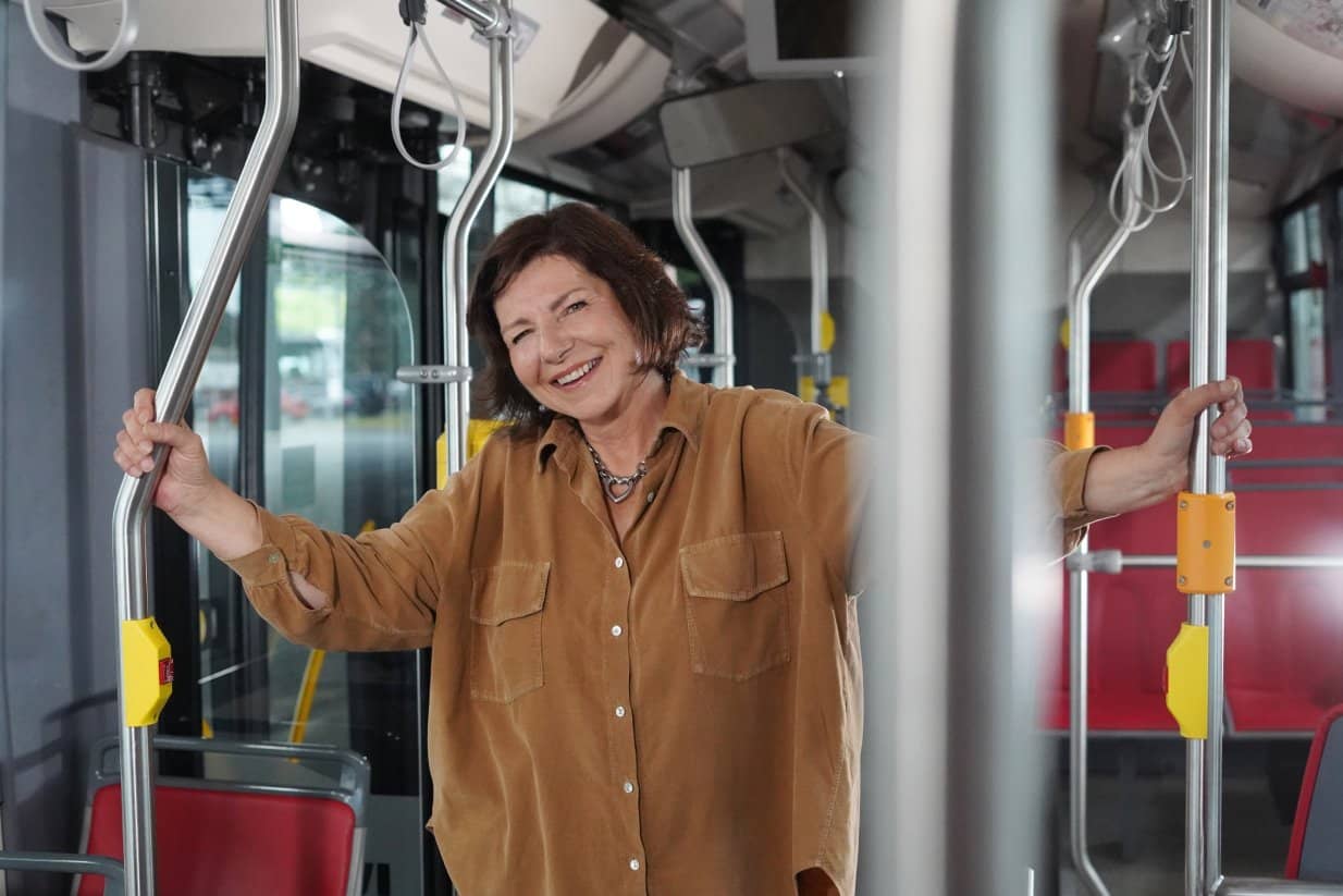 Usmívající se hnědovlasá žena v hořčicově žluté košili pózuje uvnitř moderního autobusu a drží se rukama svislých kovových tyčí pro cestující.