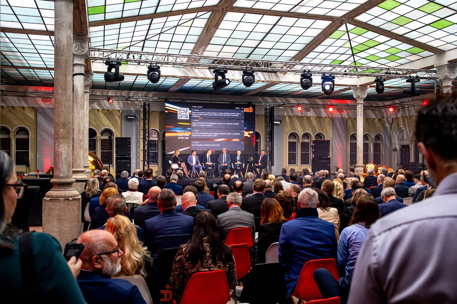 Konference s panelovou diskusí v zaplněném historickém sále s proskleným stropem. Na pódiu v pozadí sedí diskutující před velkou obrazovkou, v popředí je publikum sedící na židlích.