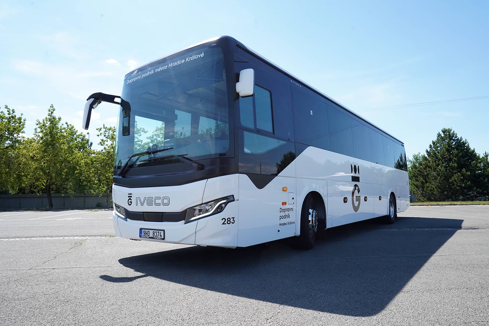 Moderní bílo-černý autokar značky Iveco s logem Dopravního podniku města Hradce Králové a evidenčním číslem 283. Autobus stojí na prázdném asfaltovém parkovišti, v pozadí je zelená vegetace a jasná modrá obloha.