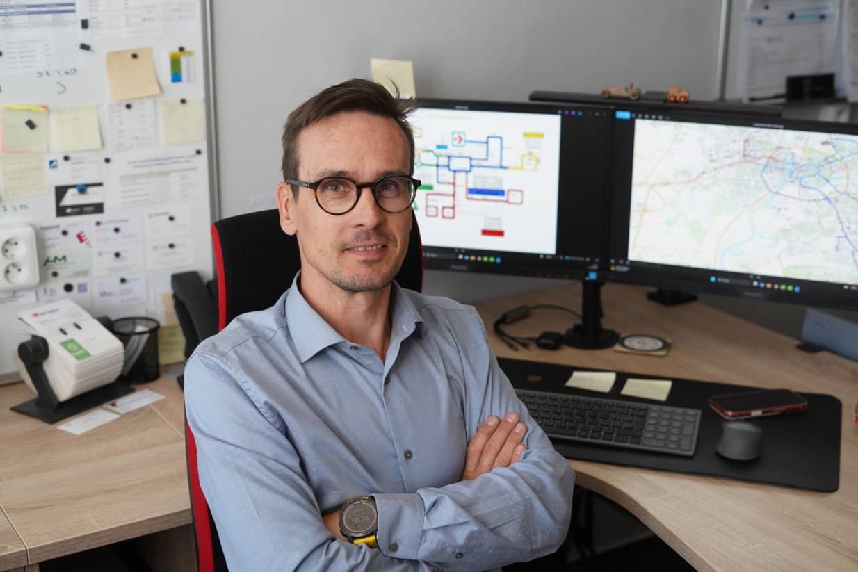 A man in glasses and a light blue shirt is sitting in an office chair at a desk with his arms crossed over his chest, looking at the camera. Behind him on the desk are two computer monitors — a block diagram is displayed on the left one, and a map on the right one. In the left background, a wallboard with notes and papers is visible.