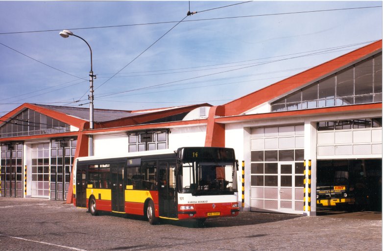 Pohled na autobus Karosa Renault Citybus (evidenční číslo 101) v červeno-žluto-bílém provedení, stojící na dlážděném nádvoří vozovny před garážovými vraty s červenou střechou. Nad areálem je vidět trolejové vedení a sloup veřejného osvětlení.