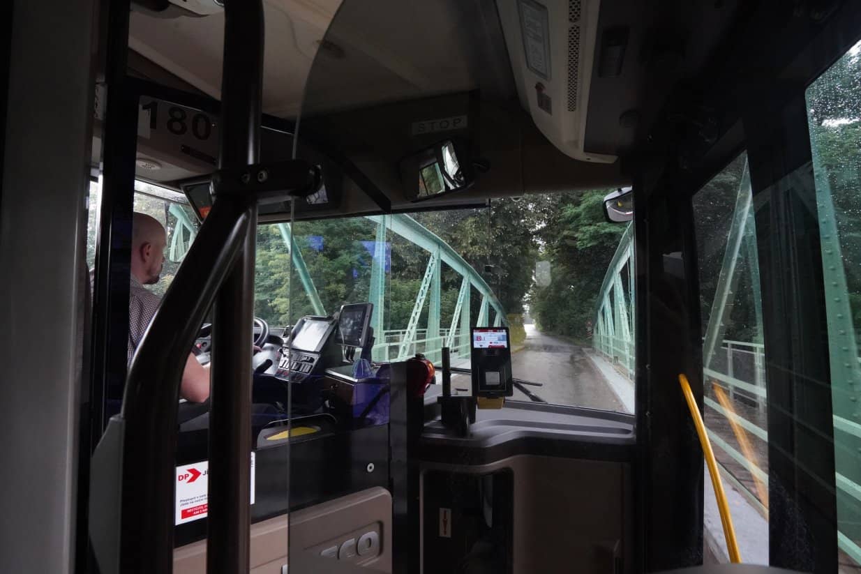Pohled z interiéru autobusu přes čelní sklo na řidiče a cestu vedoucí přes úzký zelený ocelový příhradový most obklopený stromy. V popředí je vidět palubní deska s odbavovacím terminálem na jízdenky a ochranná skleněná zástěna kabiny řidiče.