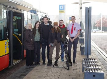 Skupina šesti lidí pózuje na autobusovém terminálu vedle moderního červeno-žlutého autobusu. Mezi nimi stojí profesionální kamera na stativu. V pozadí je vidět označník s číslem terminálu 13 a budovy v mlhavém oparu. Lidé jsou oblečeni v civilním oblečení (zimní bundy, čepice), jeden muž na pravém kraji má na sobě uniformu řidiče – košili s červenou kravatou.
