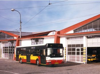 Pohled na autobus Karosa Renault Citybus (evidenční číslo 101) v červeno-žluto-bílém provedení, stojící na dlážděném nádvoří vozovny před garážovými vraty s červenou střechou. Nad areálem je vidět trolejové vedení a sloup veřejného osvětlení.