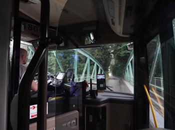 Pohled z interiéru autobusu přes čelní sklo na řidiče a cestu vedoucí přes úzký zelený ocelový příhradový most obklopený stromy. V popředí je vidět palubní deska s odbavovacím terminálem na jízdenky a ochranná skleněná zástěna kabiny řidiče.
