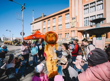 Maskot lva Gustíka baví rodiče s dětmi na prostranství před budovou Muzea východní Čech a fontánou, v pozadí stojí žlutočerný turistický vláček.