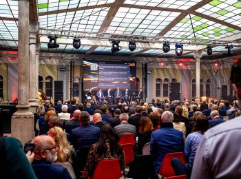 Konference s panelovou diskusí v zaplněném historickém sále s proskleným stropem. Na pódiu v pozadí sedí diskutující před velkou obrazovkou, v popředí je publikum sedící na židlích.